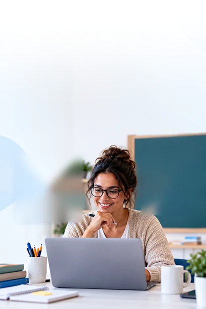 Professora sorrindo diante do notebook, em uma mesa com livros, canetas e uma x�cara.